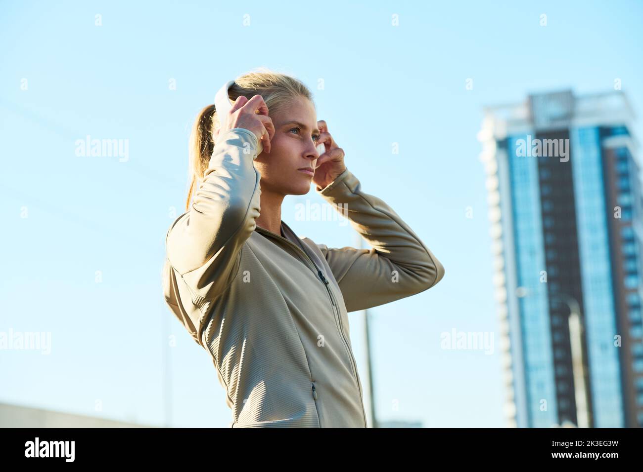 Side view of young blond woman in grey sport jacket putting headphones on her head while standing against blue sky and skyscraper Stock Photo