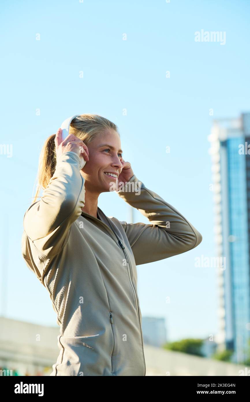 Side view of young smiling sportswoman listening to music in headphones while standing in front of camera in the city Stock Photo