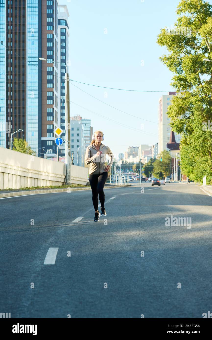 Young sportswoman in activewear running along asphalt road in the morning against modern buildings and blue sky Stock Photo