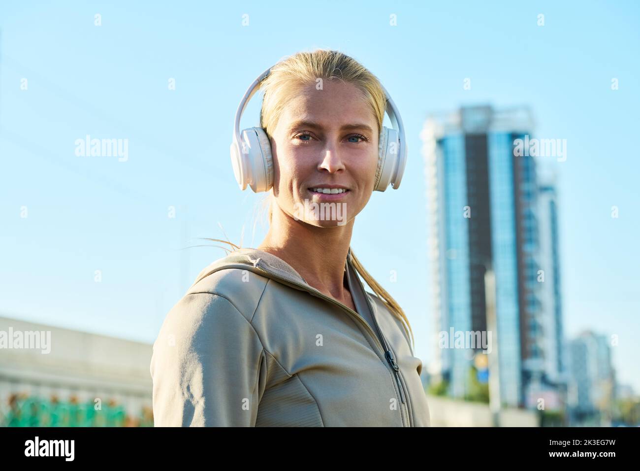 Happy young blond woman in white leather headphones and grey sport jacket looking at camera in urban environment after workout Stock Photo