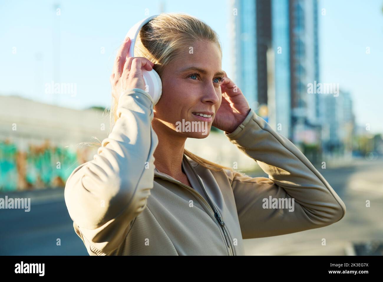 Side view of happy young blond sportswoman touching headphones on her head while listening to music in urban environment Stock Photo