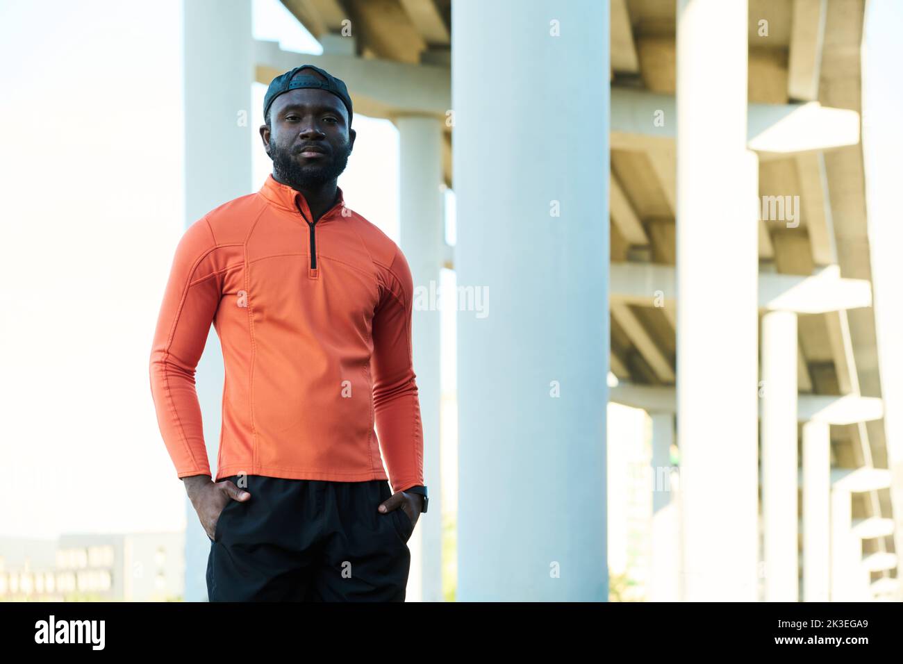 Young African American sportsman in red sport jacket looking at camera while standing against part of architectural structure outdoors Stock Photo