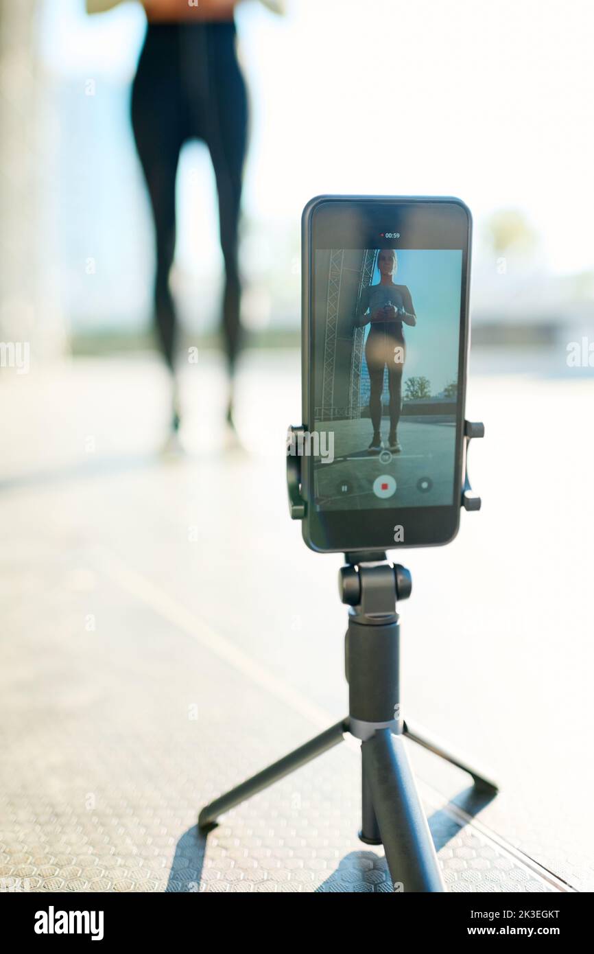 Tripod with smartphone during video record of sportswoman having outdoor workout while standing in urban environment Stock Photo