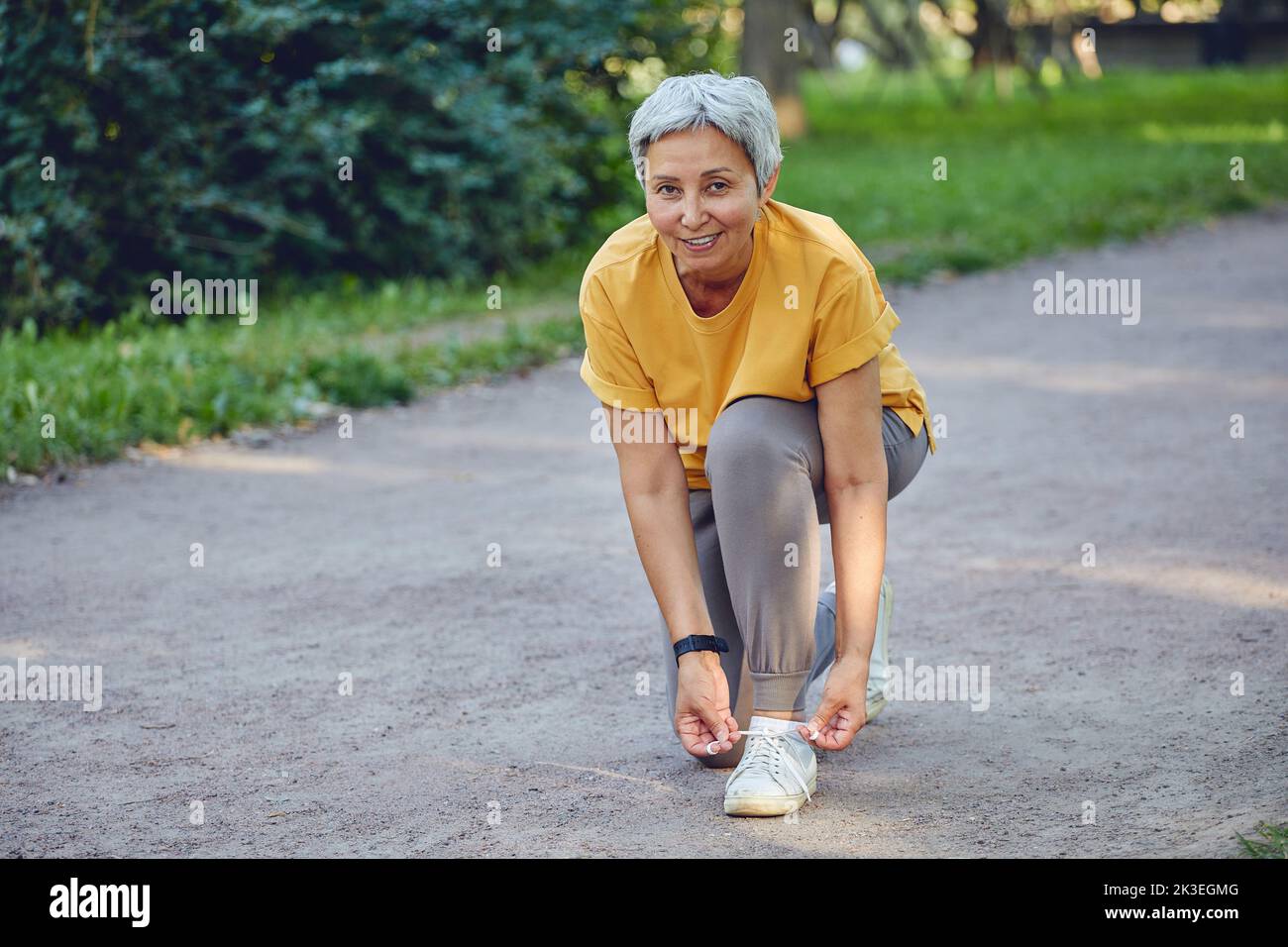 Elderly middle-aged Asia woman in sportive wear ties shoelaces ready for morning jogging or sportive walk stroll in summer park smile look at camera. Stock Photo