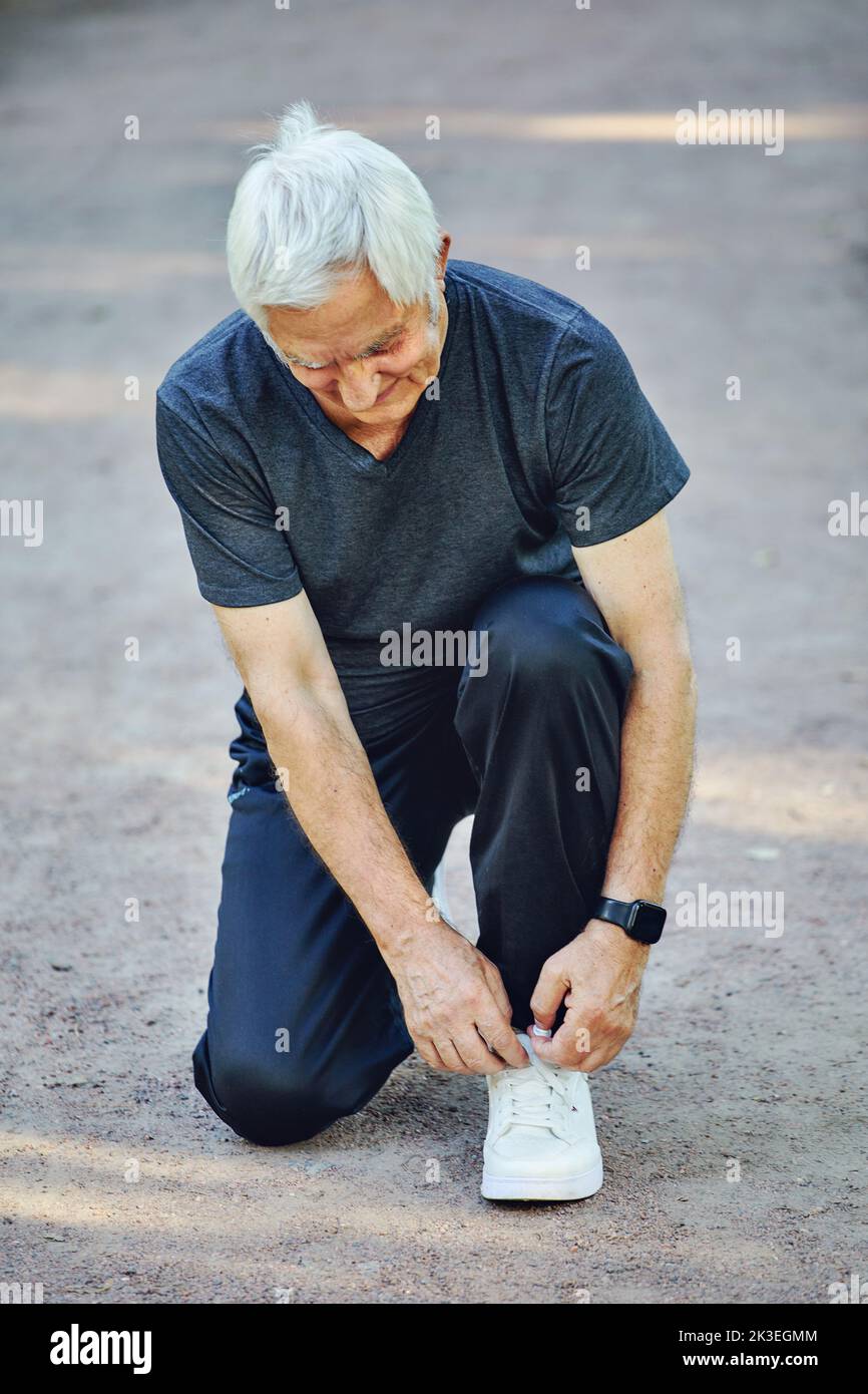 Elderly Caucasian man in sportive wear ties shoelaces ready for morning jogging or sportive walk stroll in summer park. Healthy active lifestyle of re Stock Photo