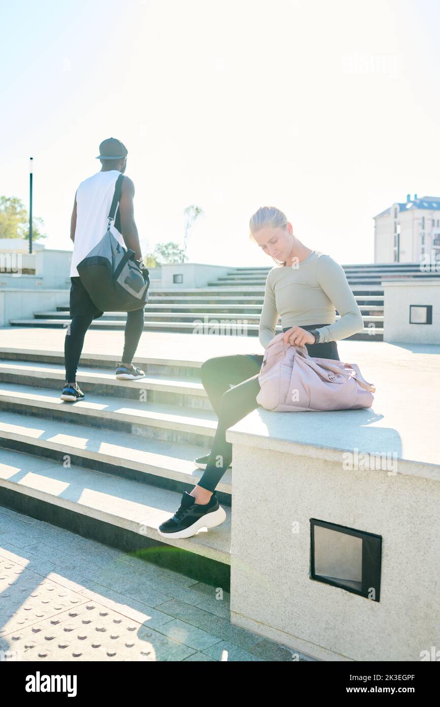 African American man with sport bag passing by blond girl sitting on concrete edge and having rest after workout or jogging Stock Photo