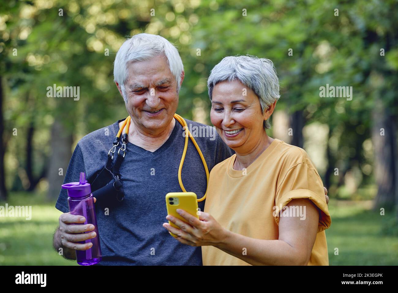 Attractive elderly grey-haired wife and husband using modern smartphone check calories counter or tracking after sportive work out in summer park. Tec Stock Photo