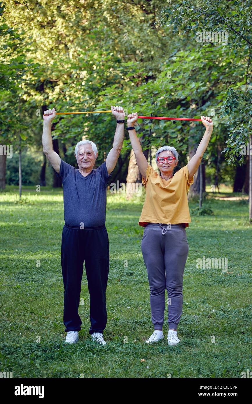 Senior spouses wear sportswear doing exercises outdoor in summer park in the morning using resistance rubber bands. Healthy lifestyle, active retired Stock Photo