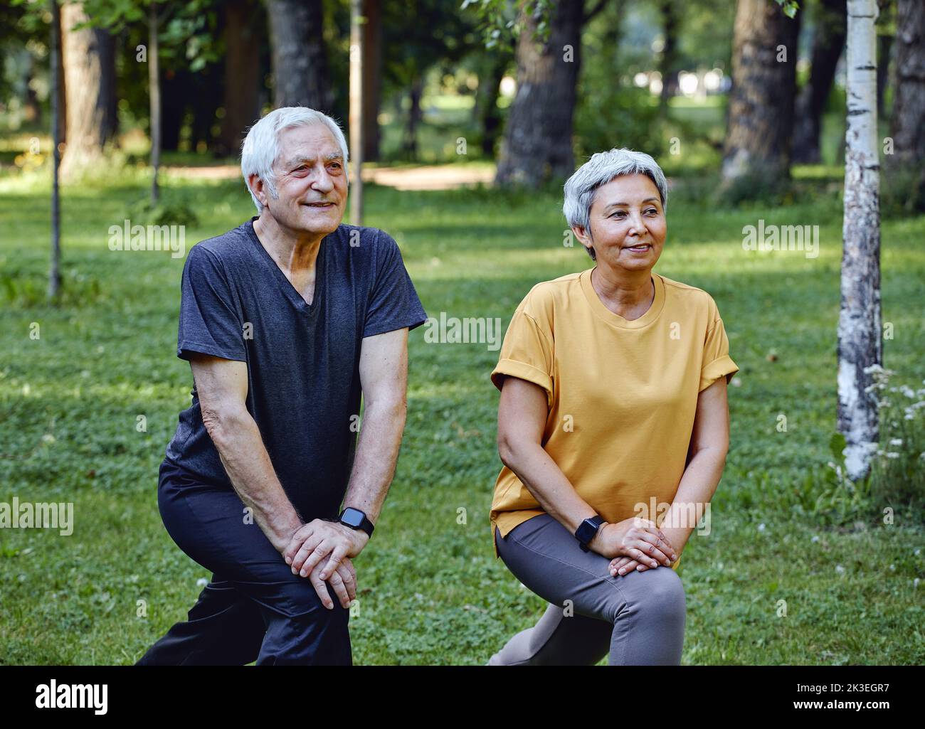 Senior spouses doing sportive exercises outdoor in summer park in the morning. Hoary healthy older couple warming-up, doing squats smile enjoy active Stock Photo