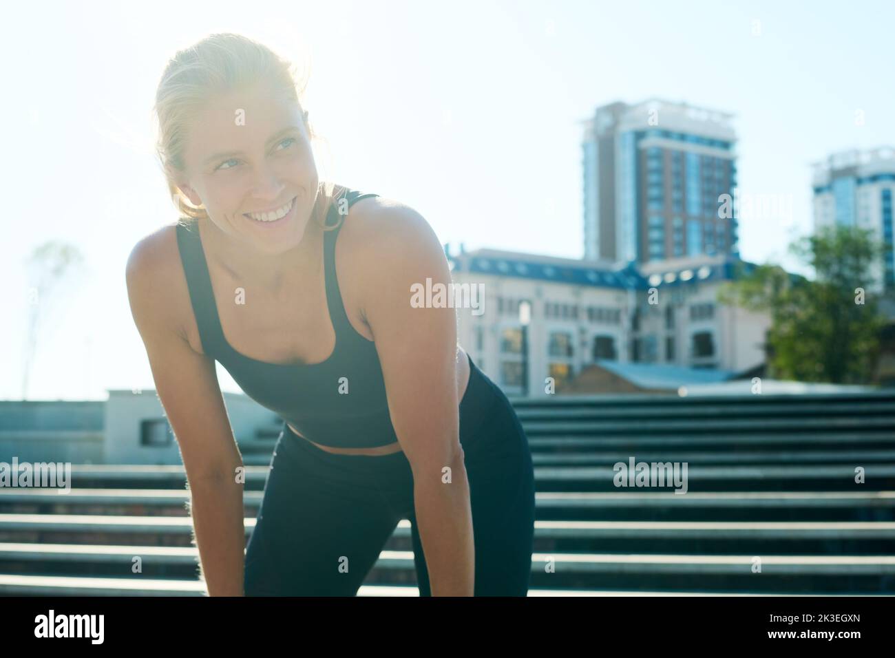 Young smiling blond female athlete in tracksuit looking aside while bending forwards in front of camera against modern architecture Stock Photo