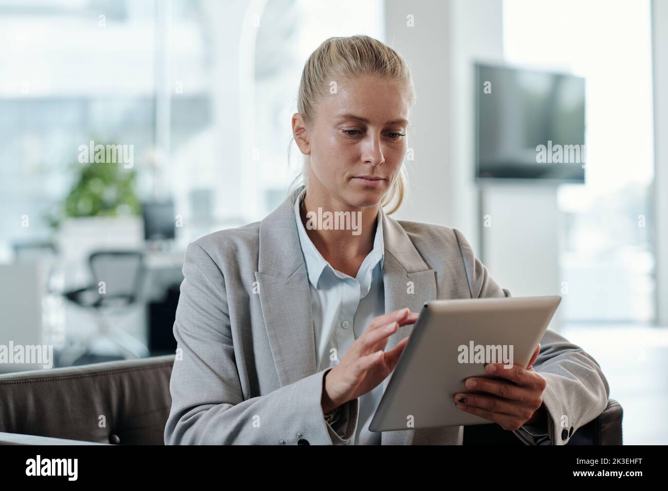 Young blond business coach in grey formalwear scrolling in tablet while sitting in armchair in office and preparing presentation for seminar Stock Photo