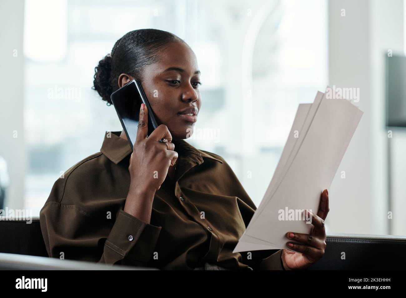 Young confident African American female manager in smart casualwear reading financial papers and talking on mobile phone in office Stock Photo