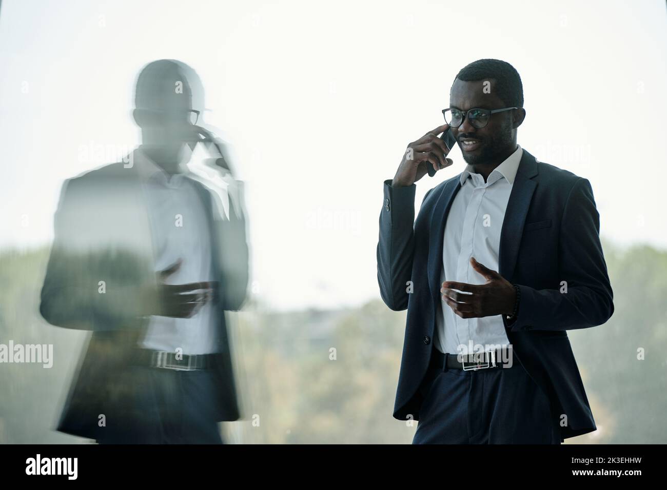 Young successful African American businessman or entrepreneur with mobile phone by ear standing in front of office window Stock Photo