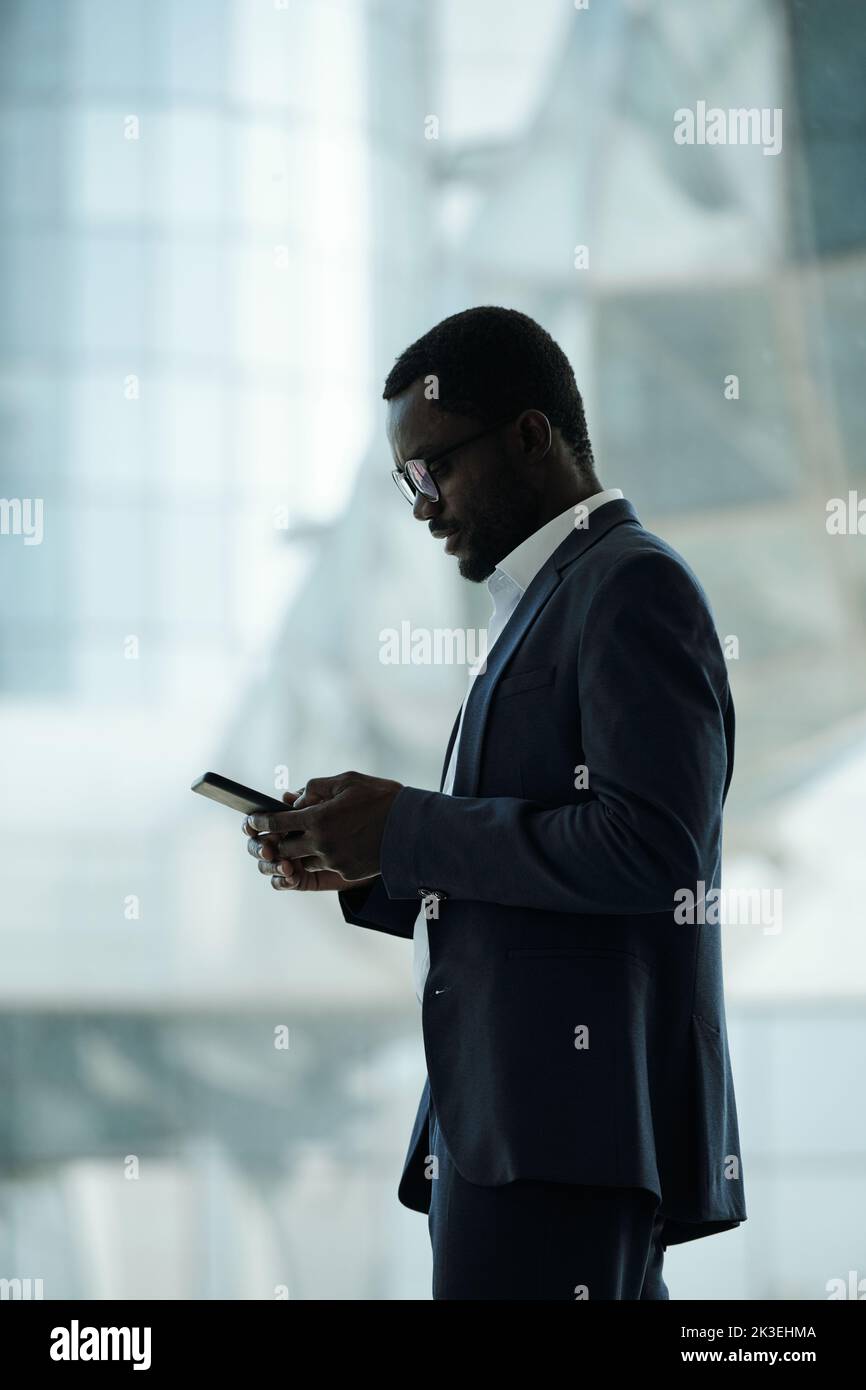 Side view of young modern businessman in elegant suit texting in smartphone while standing in front of camera against window Stock Photo