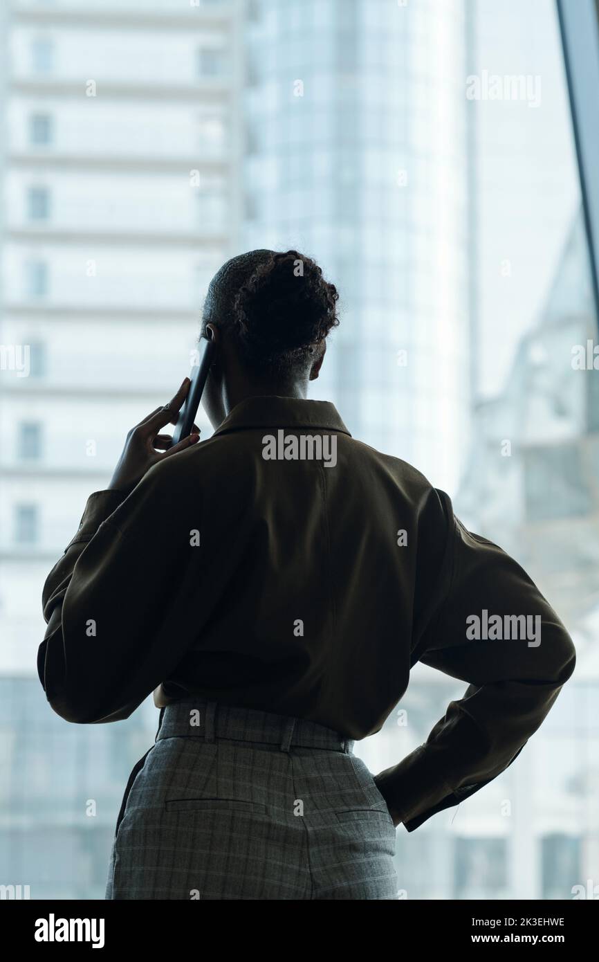 Rear view of young black woman with hair bun standing in front of large office window and communicating on mobile phone Stock Photo