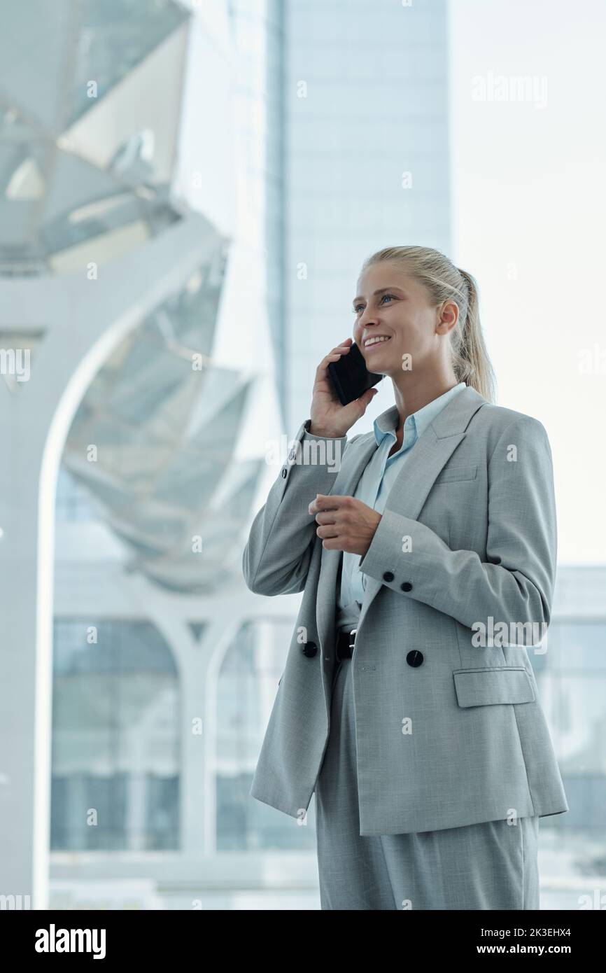 Happy young blond businesswoman in grey suit talking on mobile phone while standing by large iwndow in modern office Stock Photo