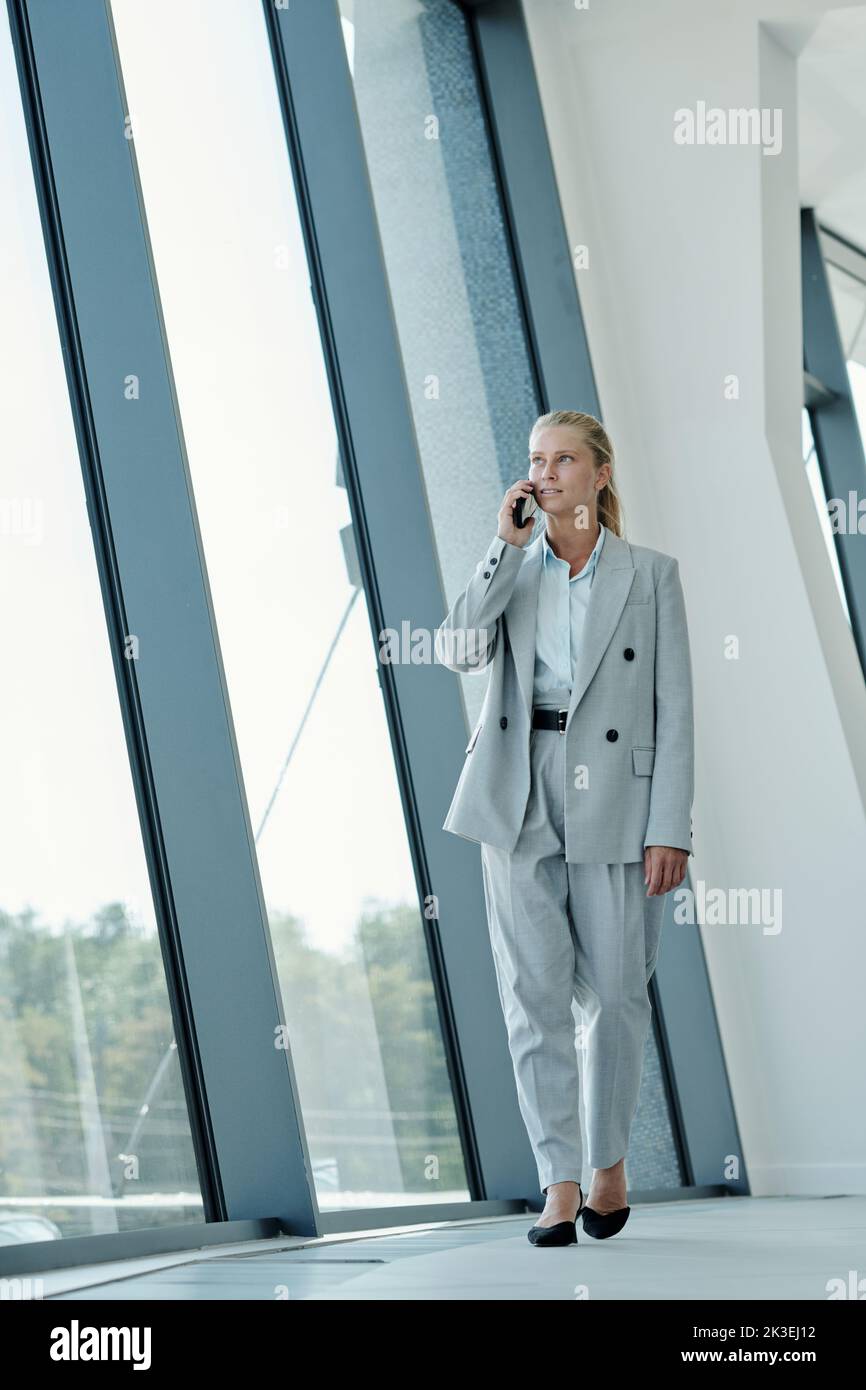 Young blond businesswoman with smartphone by ear walking along large window in corridor of modern office center and talking to client Stock Photo