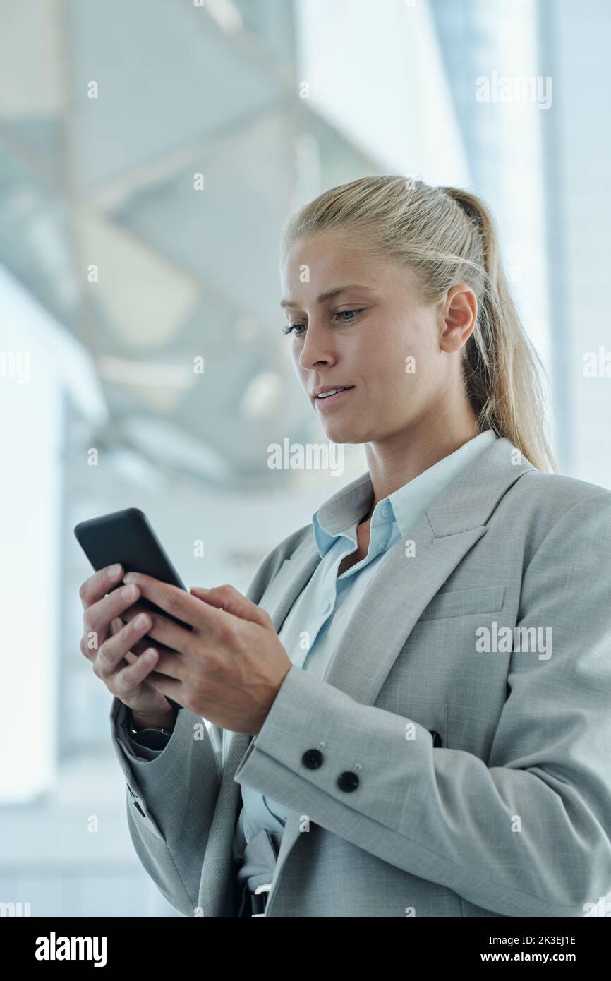 Confident blond businesswoman looking at smartphone screen during online communication while standing in front of camera in office Stock Photo