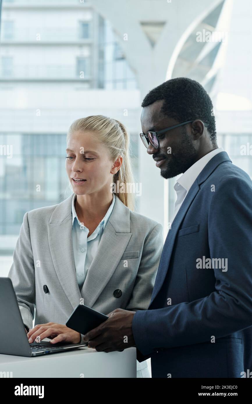 Young confident female broker in elegant grey suit discussing online data with colleague while both looking at laptop screen at meeting Stock Photo