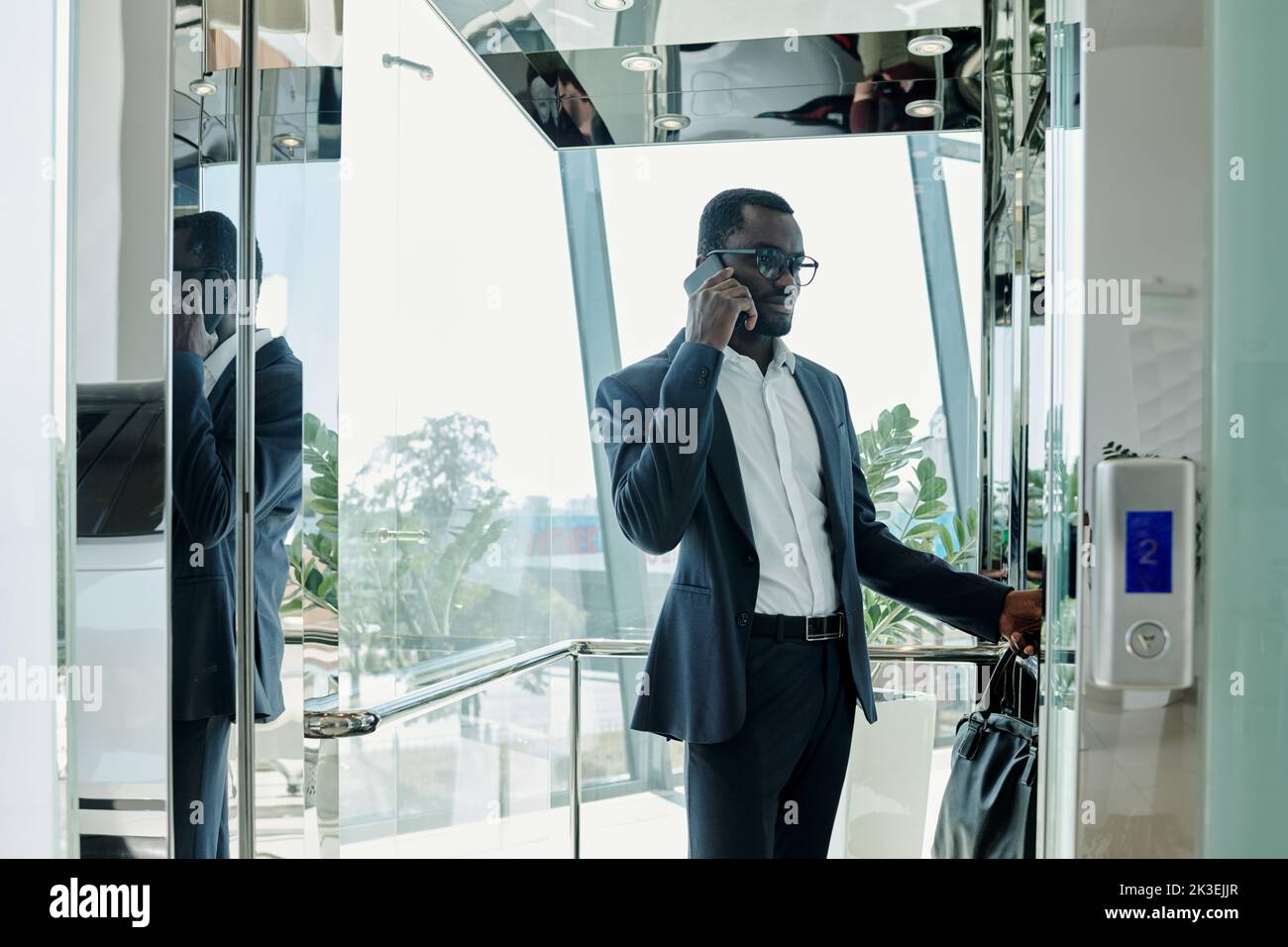 Young elegant businessman in suit standing inside transparent elevator in modern office center and communicating on mobile phone Stock Photo