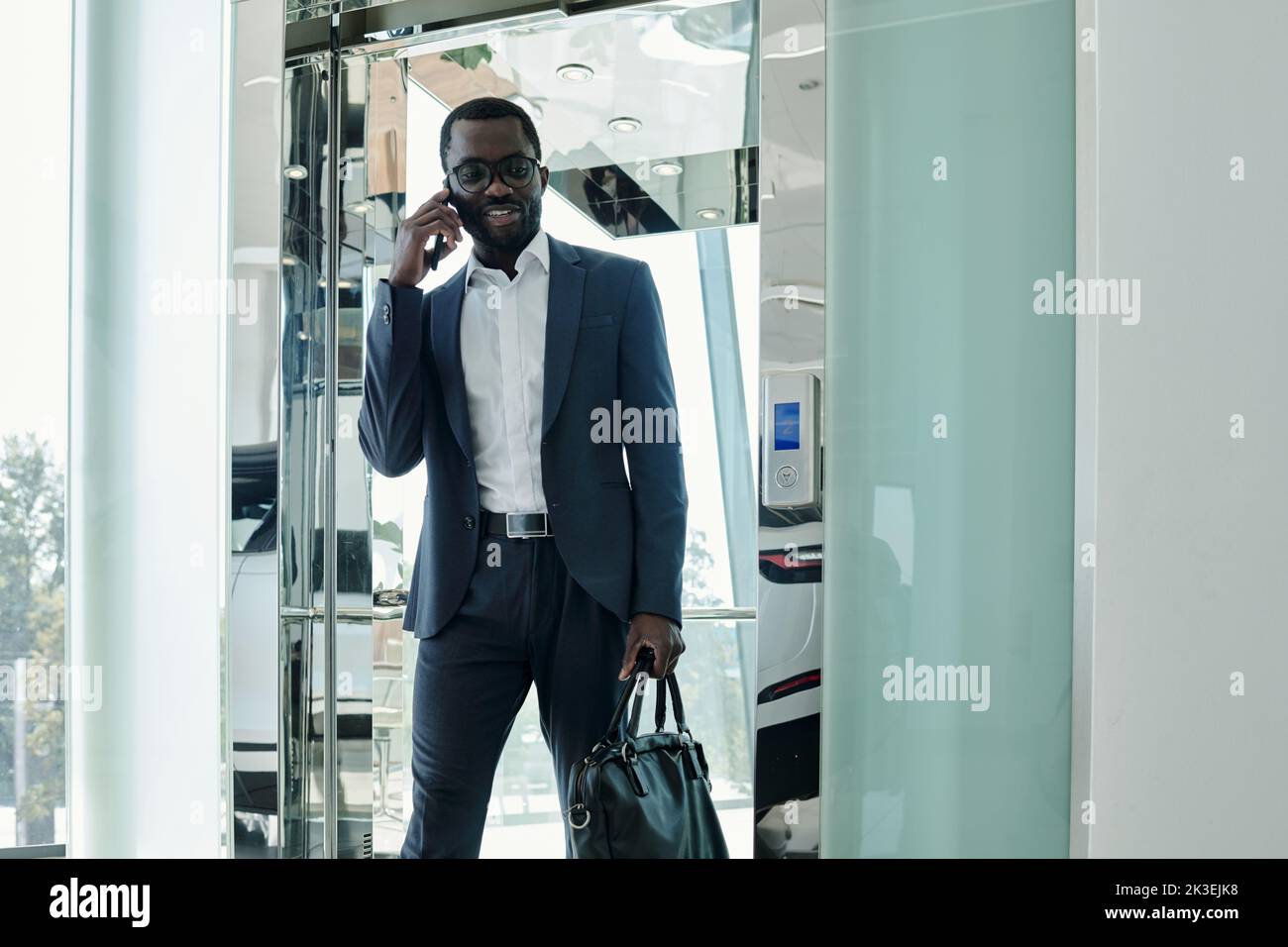 Confident African American businessman in elegant suit talking on smartphone while going out of elevator with transparent walls Stock Photo