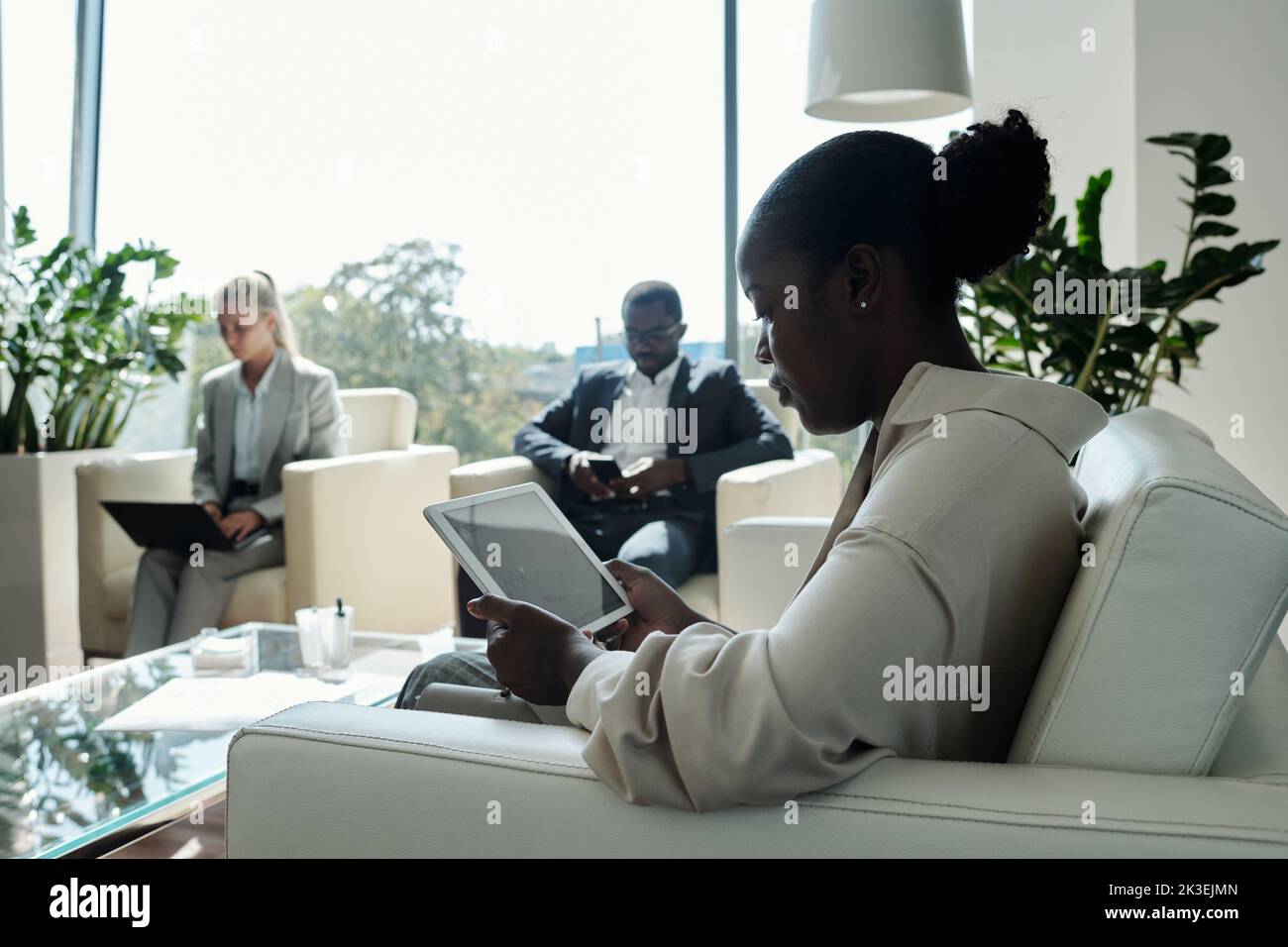 Side view of young serious female office worker with tablet sitting on white leather couch in openspace office against two coworkers Stock Photo