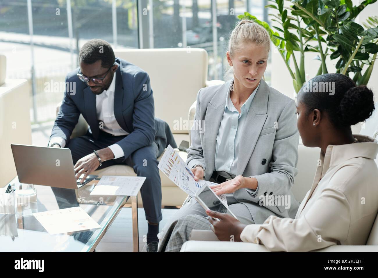 Young confident blond female economist showing document with financial data to African American colleague with tablet Stock Photo
