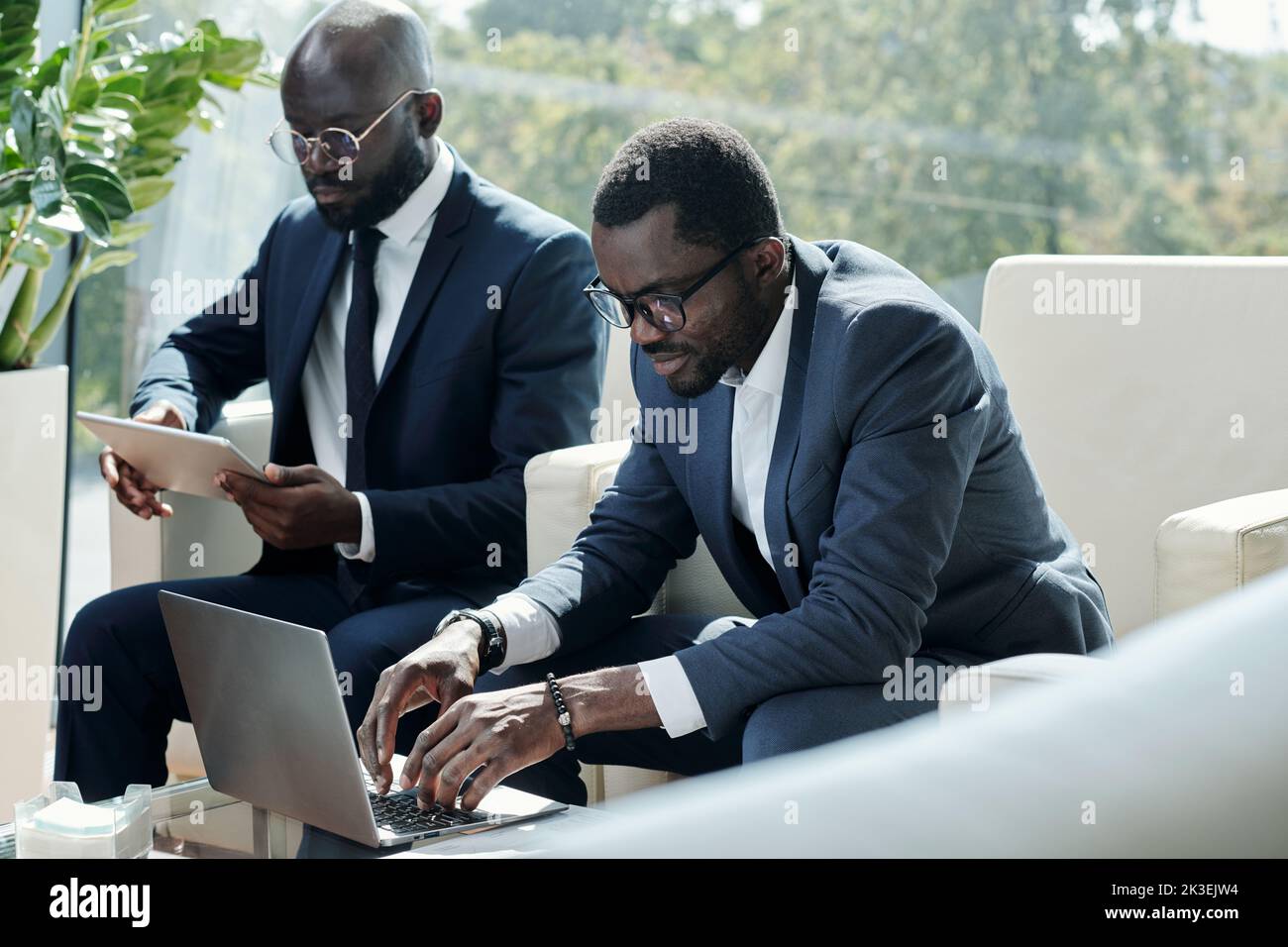 Young serious black man in formalwear typing on laptop keypad while sitting in armchair against male coworker using tablet Stock Photo