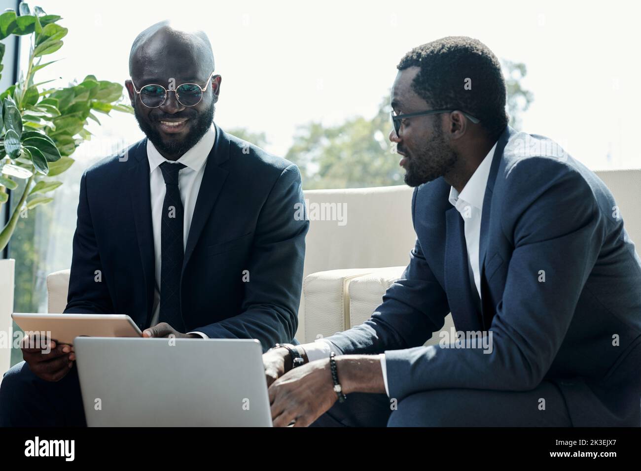Young smiling African American businessman with tablet and his colleague having discussion of project points at meeting Stock Photo