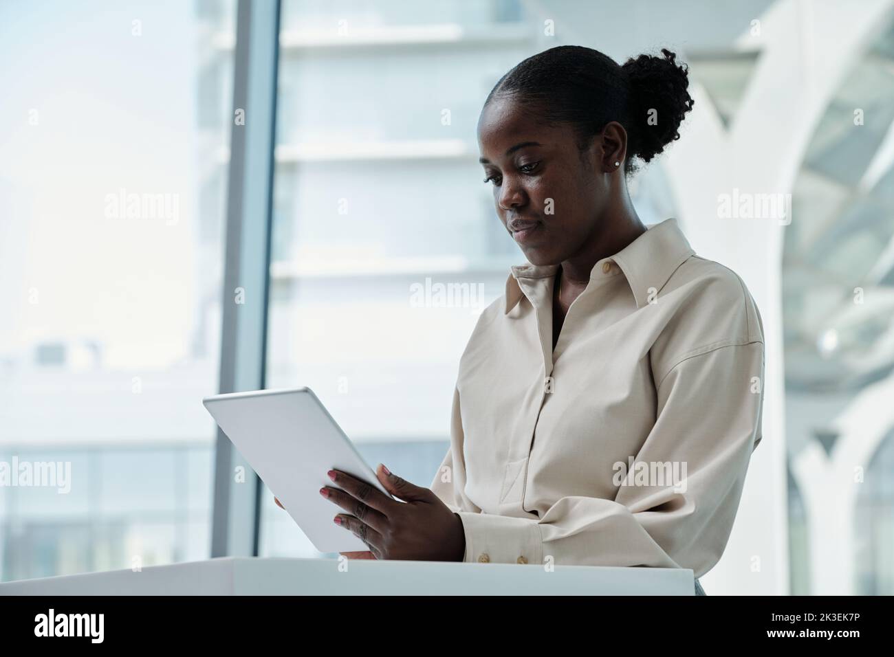 Young serious African American female analyst with tablet sitting in office against large window and looking through online data Stock Photo
