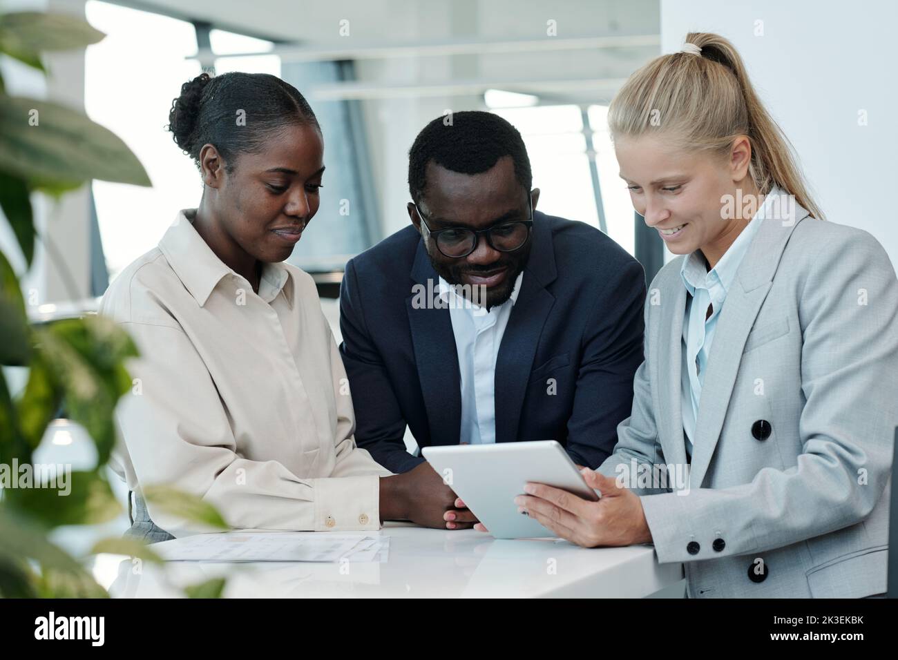 Group of young intercultural managers in formalwear looking at tablet screen while discussing financial data at working meeting Stock Photo