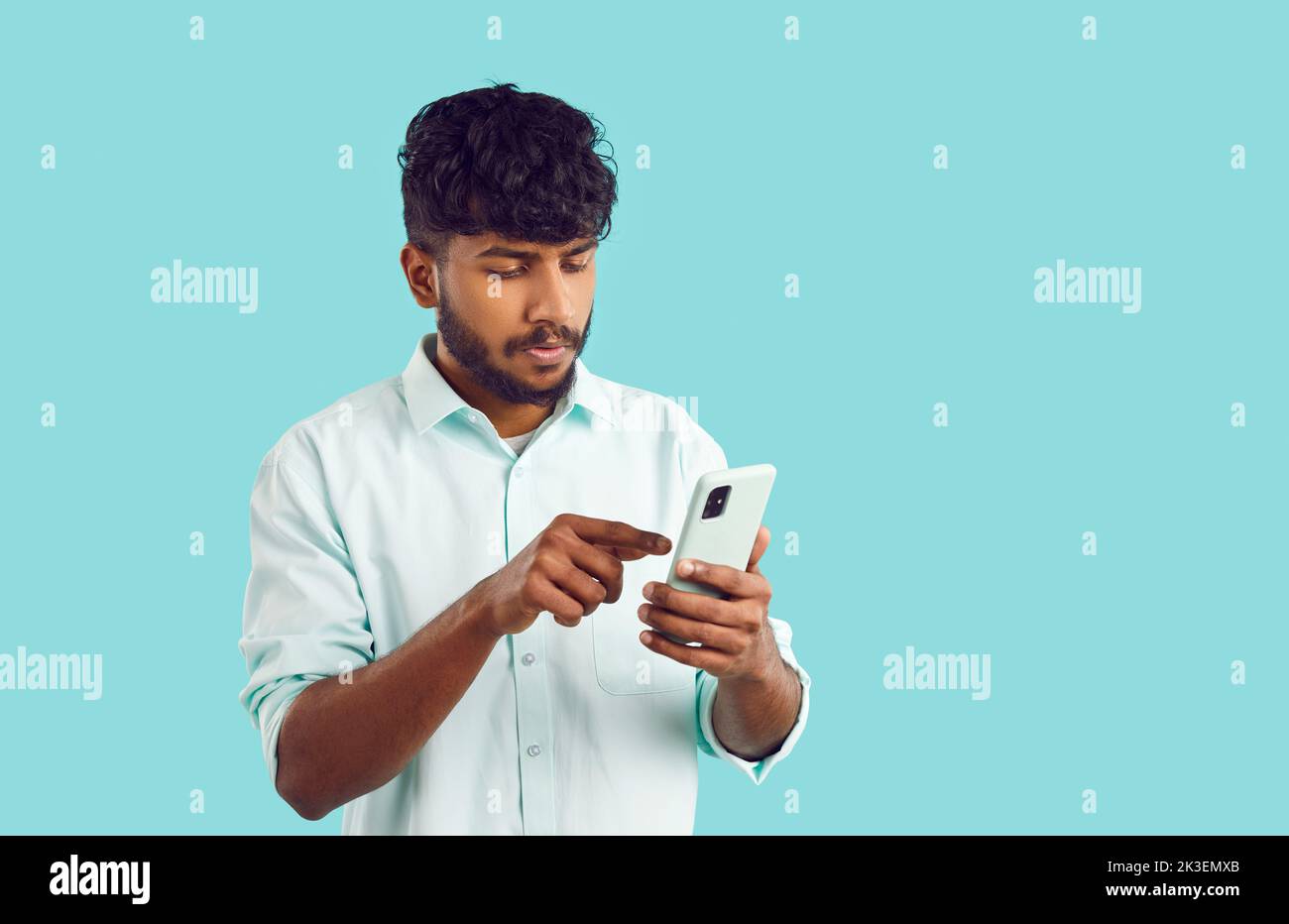 Young focused Indian man holding mobile phone reading latest news stands on turquoise background Stock Photo