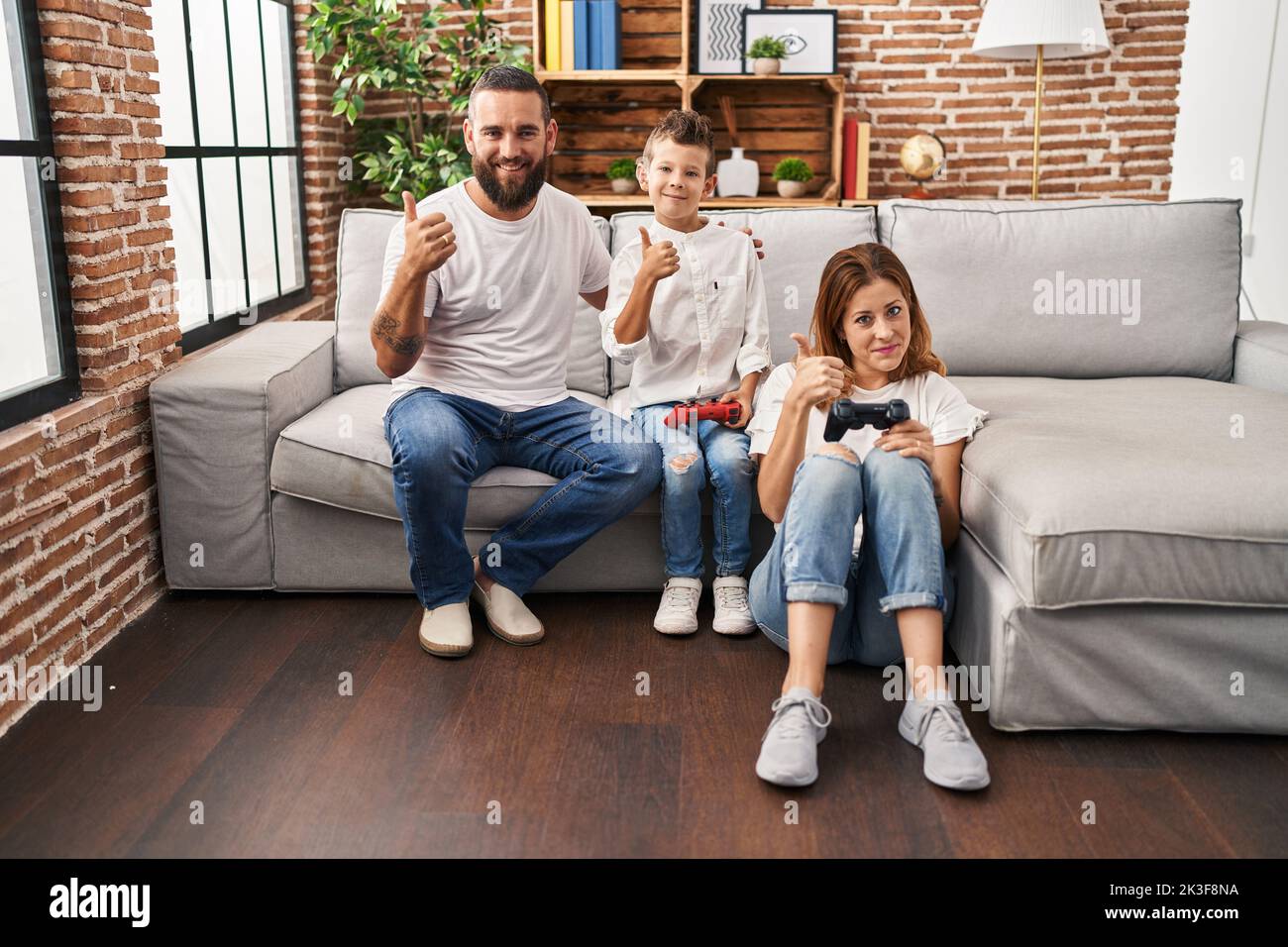 Family of three playing video game sitting on the sofa smiling happy and positive, thumb up doing excellent and approval sign Stock Photo