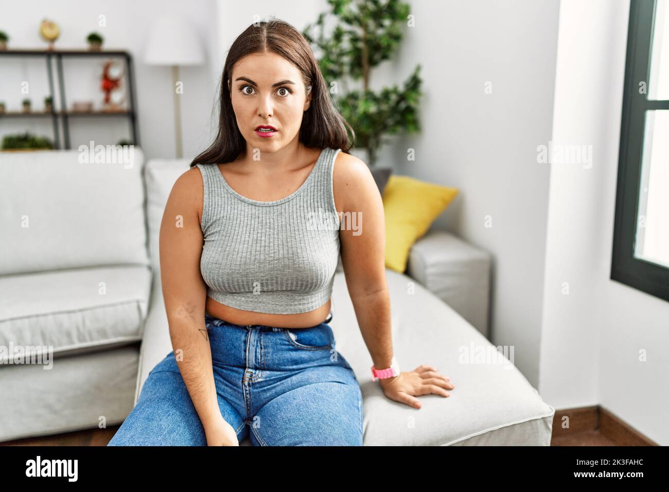 Young brunette woman sitting on the sofa at home in shock face, looking ...