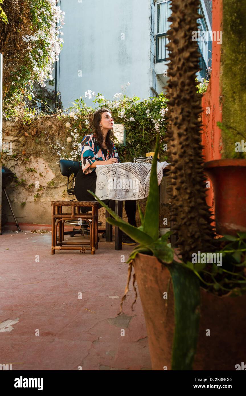vertical image of french adult woman sitting on the terrace of home studying and memorizing concepts, with a plant in the foreground and copy space. Stock Photo