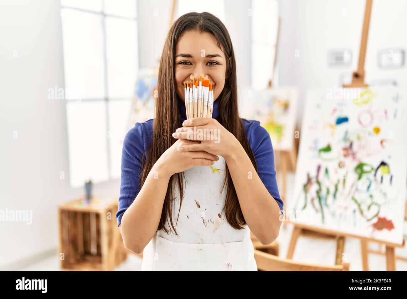 Young latin woman covering mouth with paintbrushes at art studio Stock ...