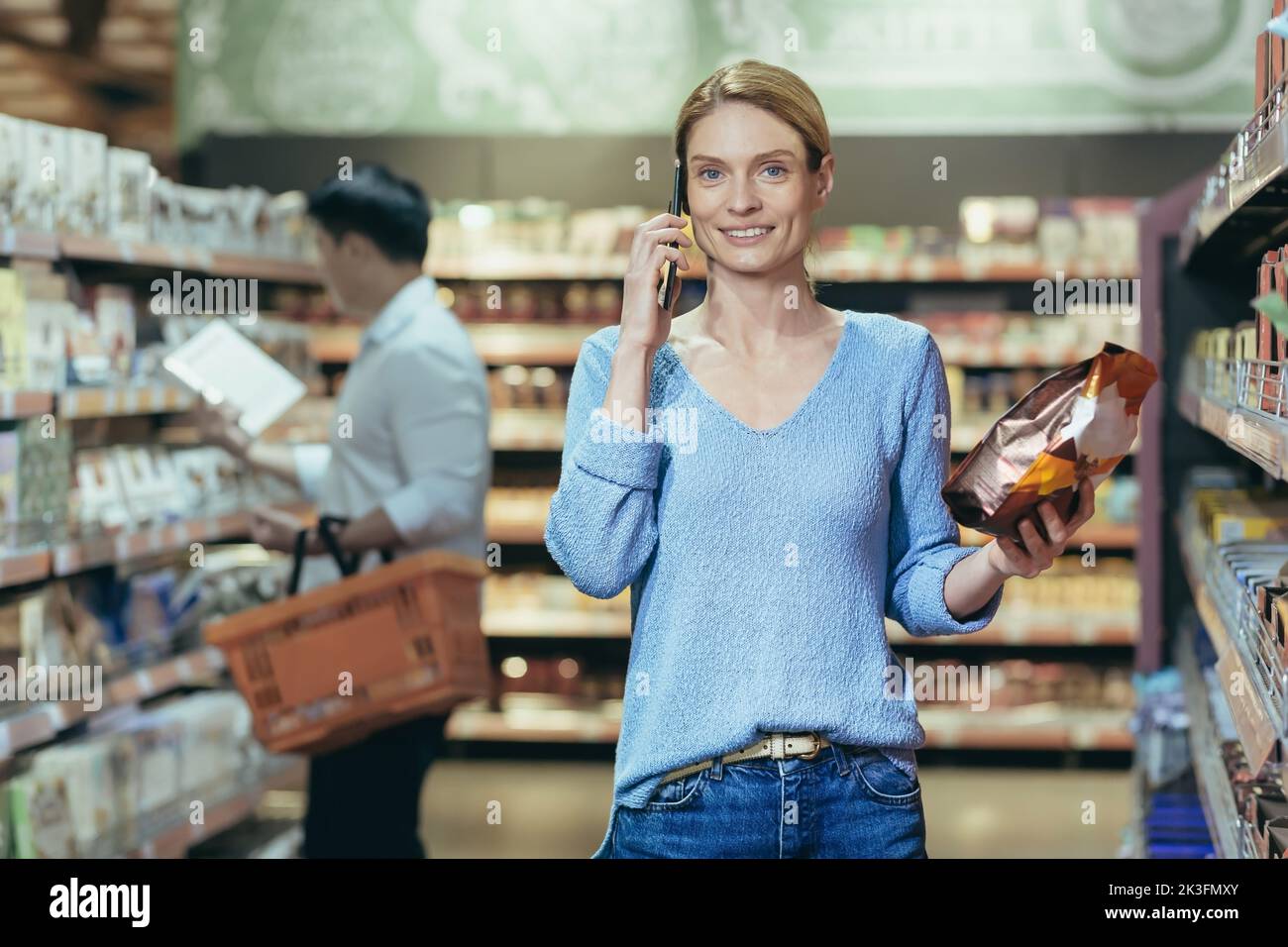 A beautiful young blonde woman is standing in a supermarket and talking on the phone, consulting, holding a pack of coffee in her hand, smiling. Shoppers in the background. Stock Photo