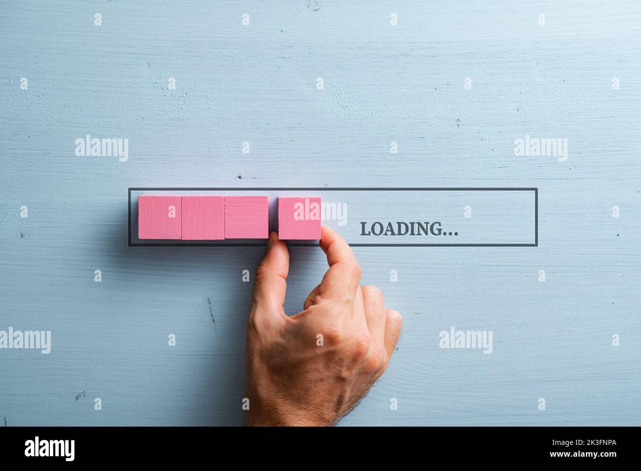 Male hand placing a pink wooden blocks into a loading bar with a Loading sign. Over pastel blue wooden background. Stock Photo