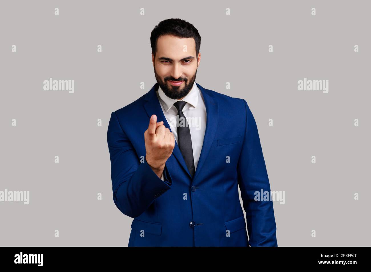 Portrait of positive smiling handsome bearded businessman calling with finger gesture, inviting to come in, wearing official style suit. Indoor studio shot isolated on gray background. Stock Photo