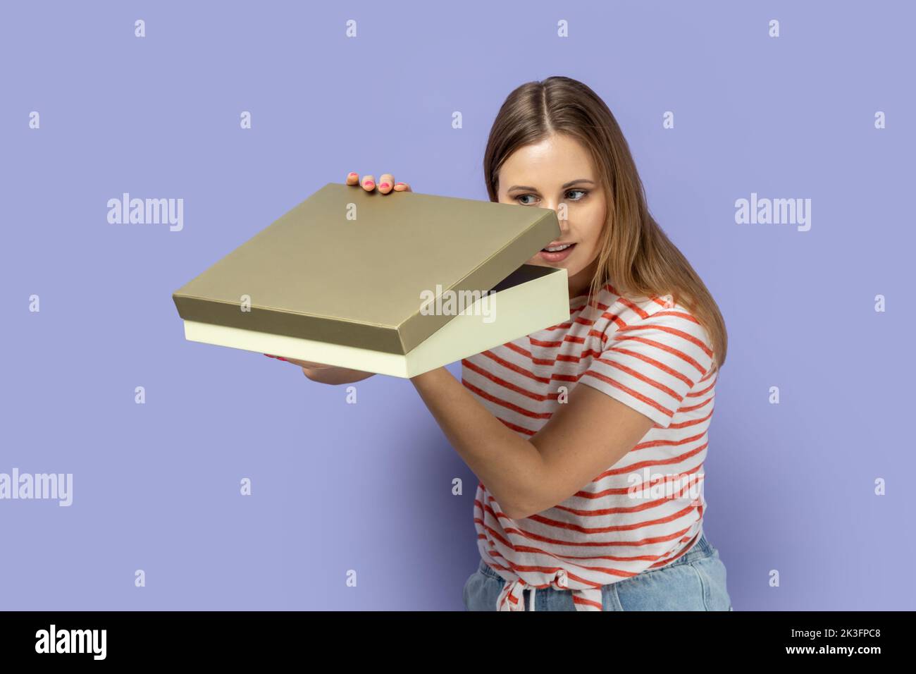 Portrait of curious calm blond woman wearing striped T-shirt standing holding present box and looking inside, interesting what inside. Indoor studio shot isolated on purple background. Stock Photo