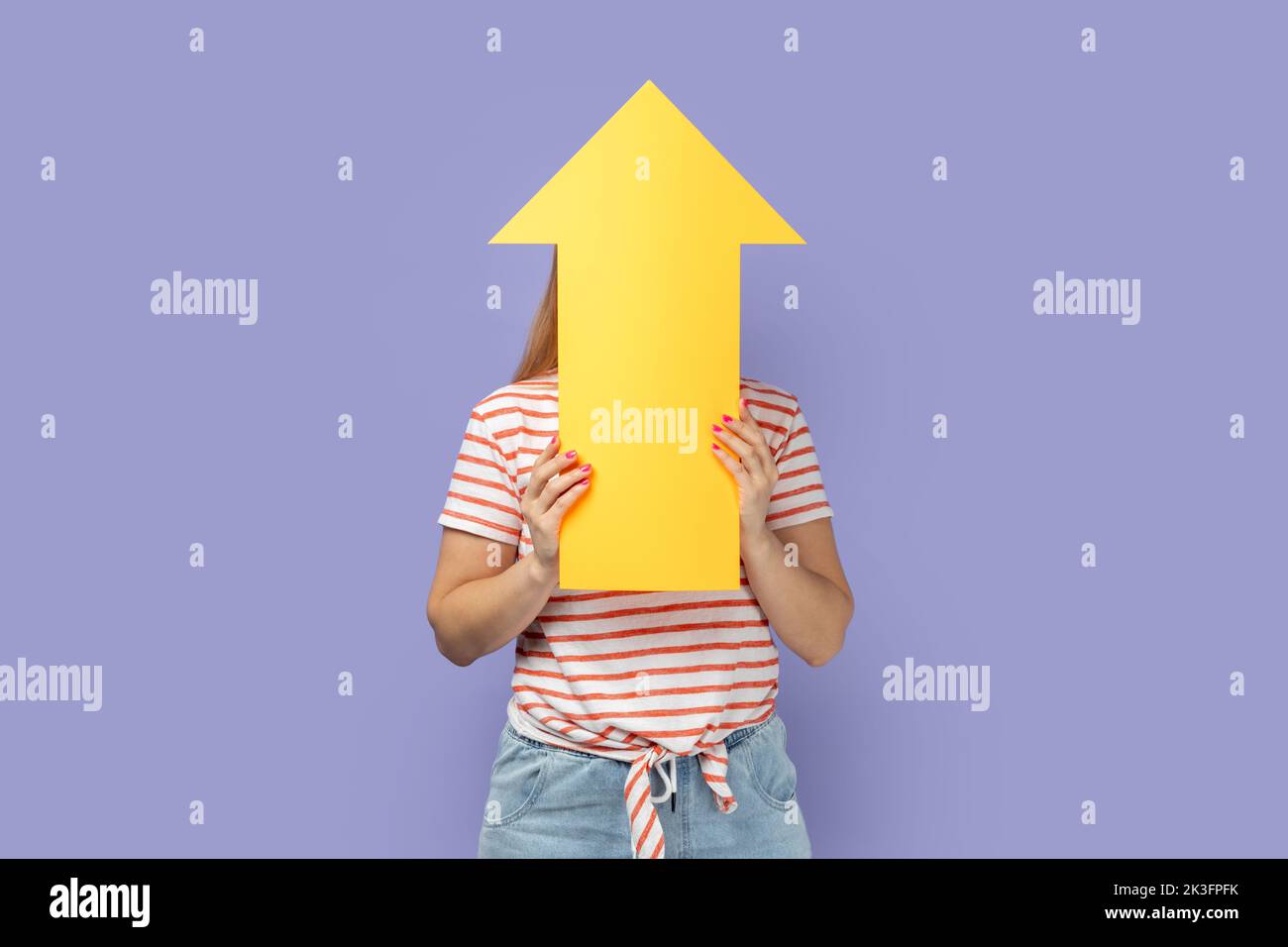 Portrait of anonymous unknown blond woman wearing striped T-shirt holding big yellow arrow pointing up, showing increase in career. Indoor studio shot isolated on purple background. Stock Photo