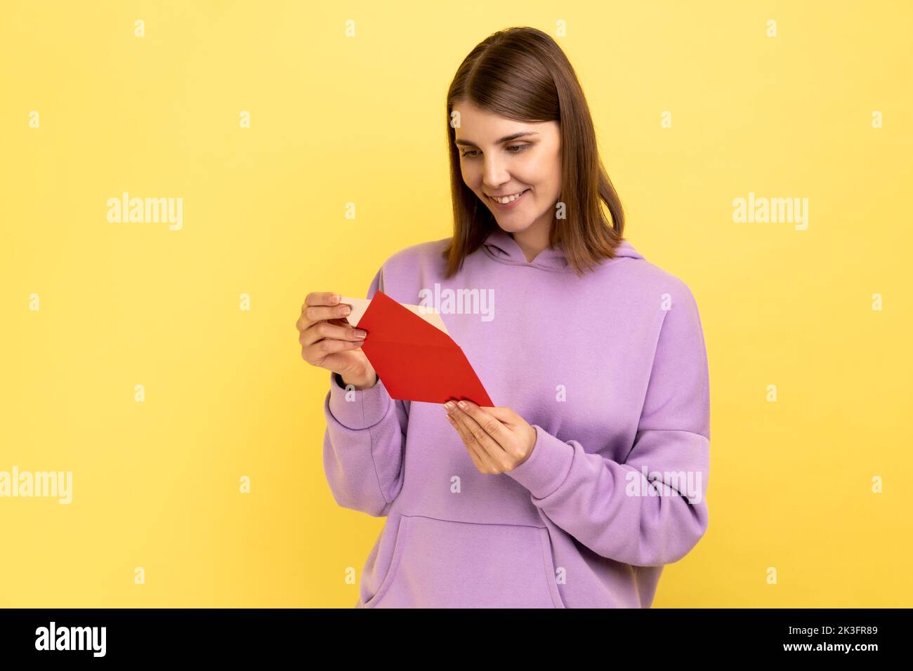 Side view of smiling young woman opening envelope and reading letter, with positive expression, got unexpected pleasant news, wearing purple hoodie. Indoor studio shot isolated on yellow background. Stock Photo