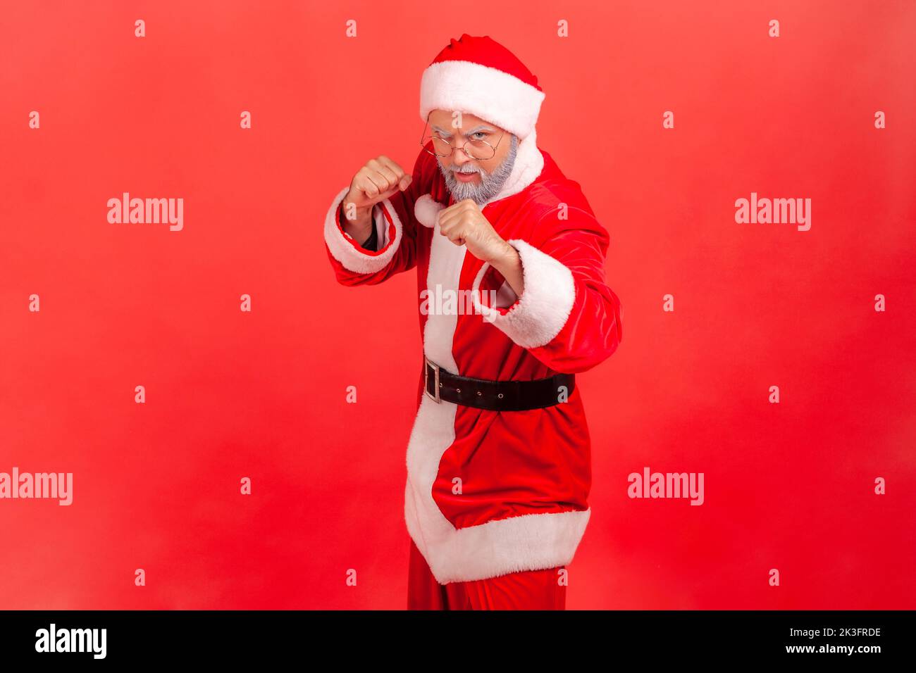 Strict elderly man with gray beard wearing santa claus costume standing with fists and being ready to fight, looking at camera with angry expression. Indoor studio shot isolated on red background. Stock Photo