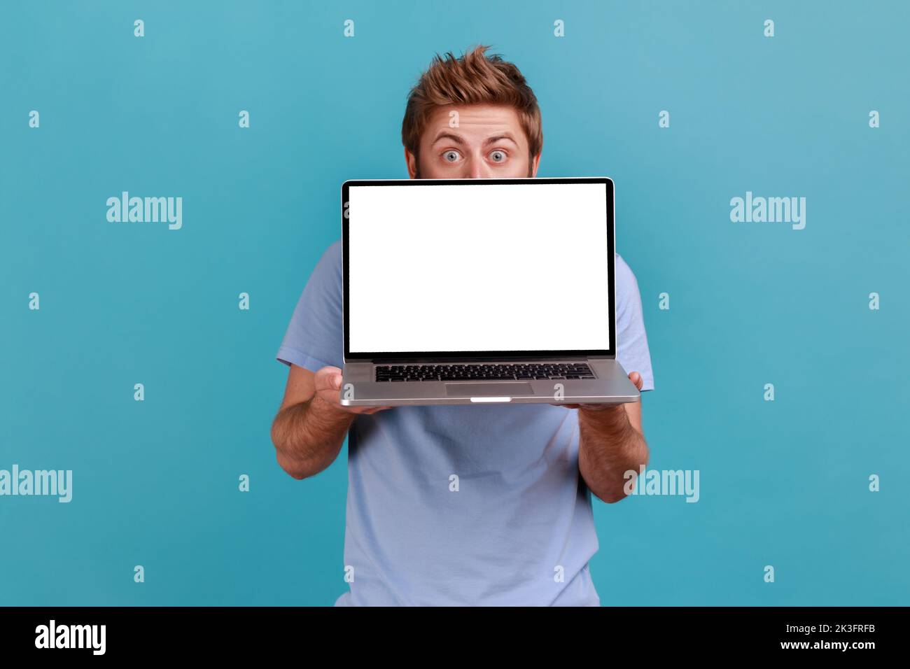 Portrait of man hiding behind white empty screen laptop, surprised with advertising area, looking at camera with big eyes, updating operation system. Indoor studio shot isolated on blue background. Stock Photo