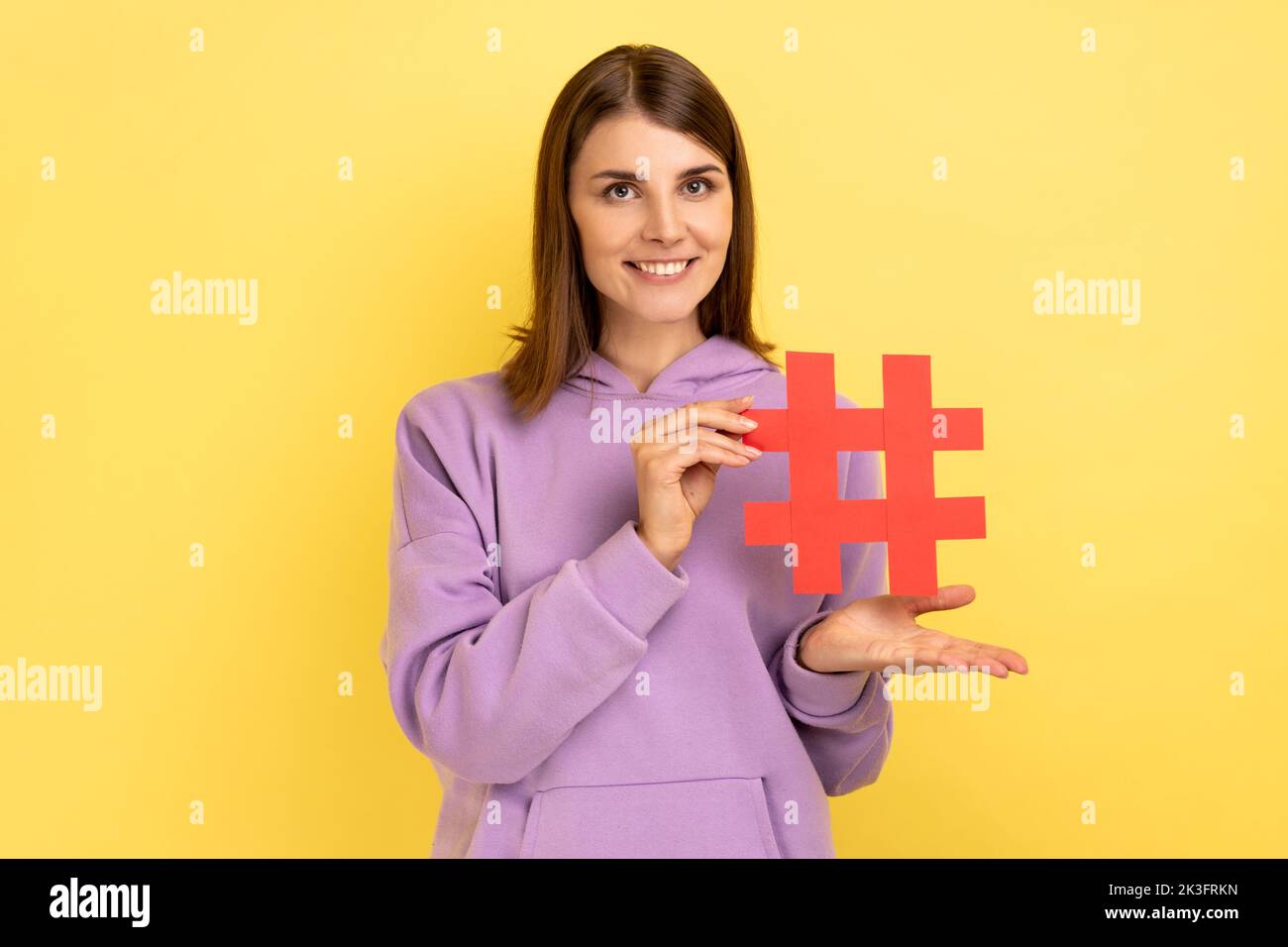 Portrait of young woman standing presenting red hashtag, tagging blog trends, viral topic in social network, wearing purple hoodie. Indoor studio shot isolated on yellow background. Stock Photo