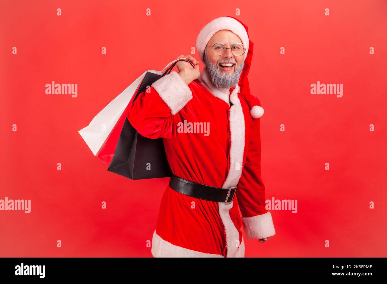 Portrait of smiling elderly man with gray beard wearing santa claus costume standing with shopping bags, buying present for winter holidays. Indoor studio shot isolated on red background. Stock Photo