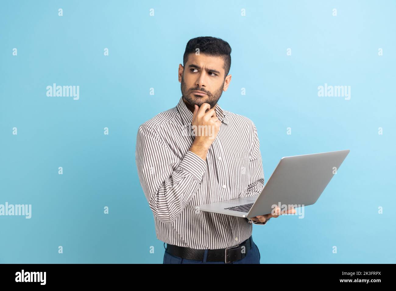 Portrait thoughtful pensive man with beard holding laptop and holding chin looking away, thinking over new business project, wearing striped shirt. Indoor studio shot isolated on blue background. Stock Photo