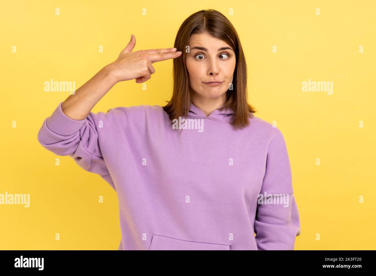 Portrait of desperate woman, pointing finger gun to head, shooting herself, making suicide gesture to stop depression, wearing purple hoodie. Indoor studio shot isolated on yellow background. Stock Photo