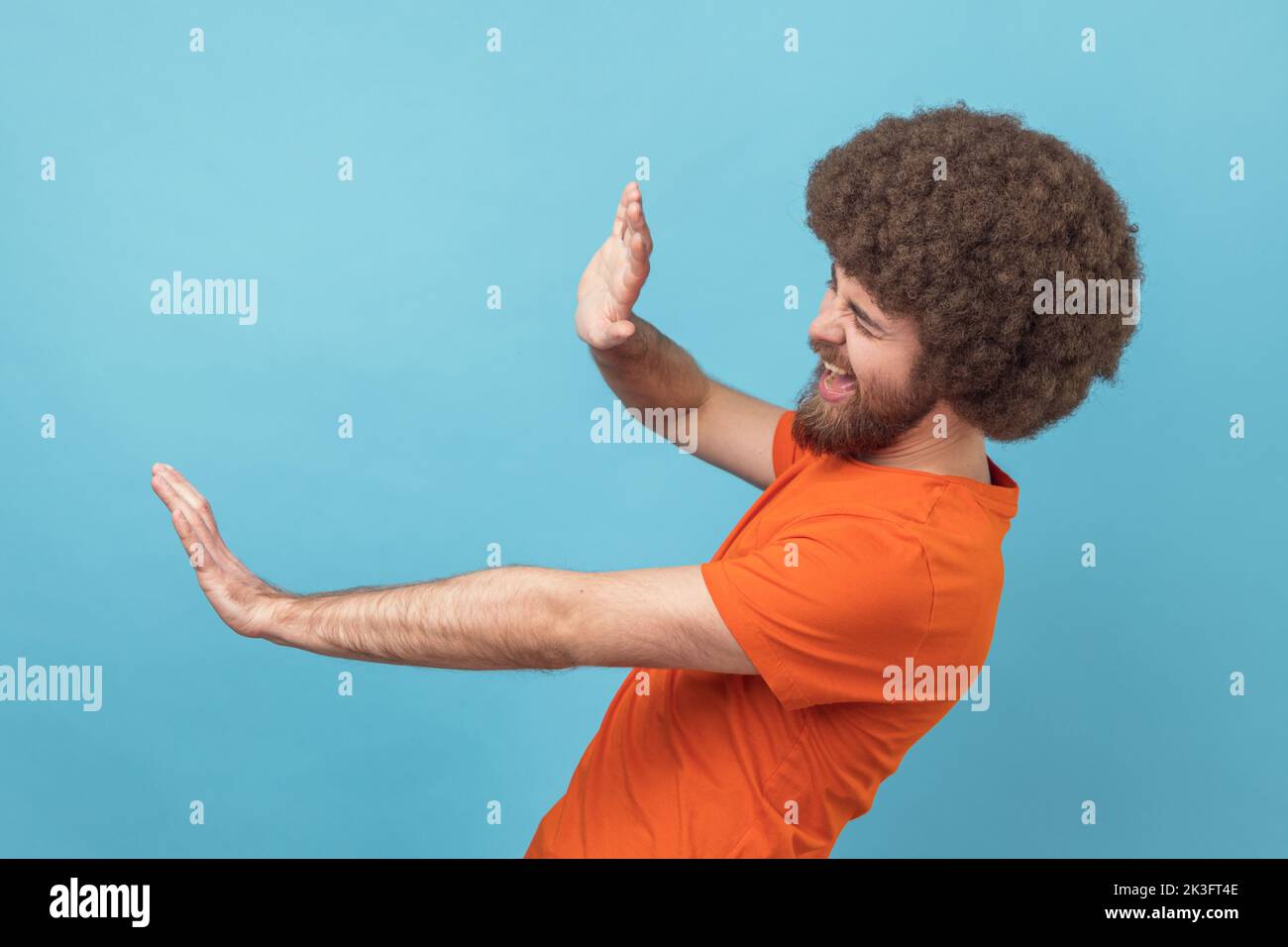 Side view of scared man with Afro hairstyle wearing orange T-shirt standing with afraid or worry face, looking ahead and blocking with his hands. Indoor studio shot isolated on blue background. Stock Photo