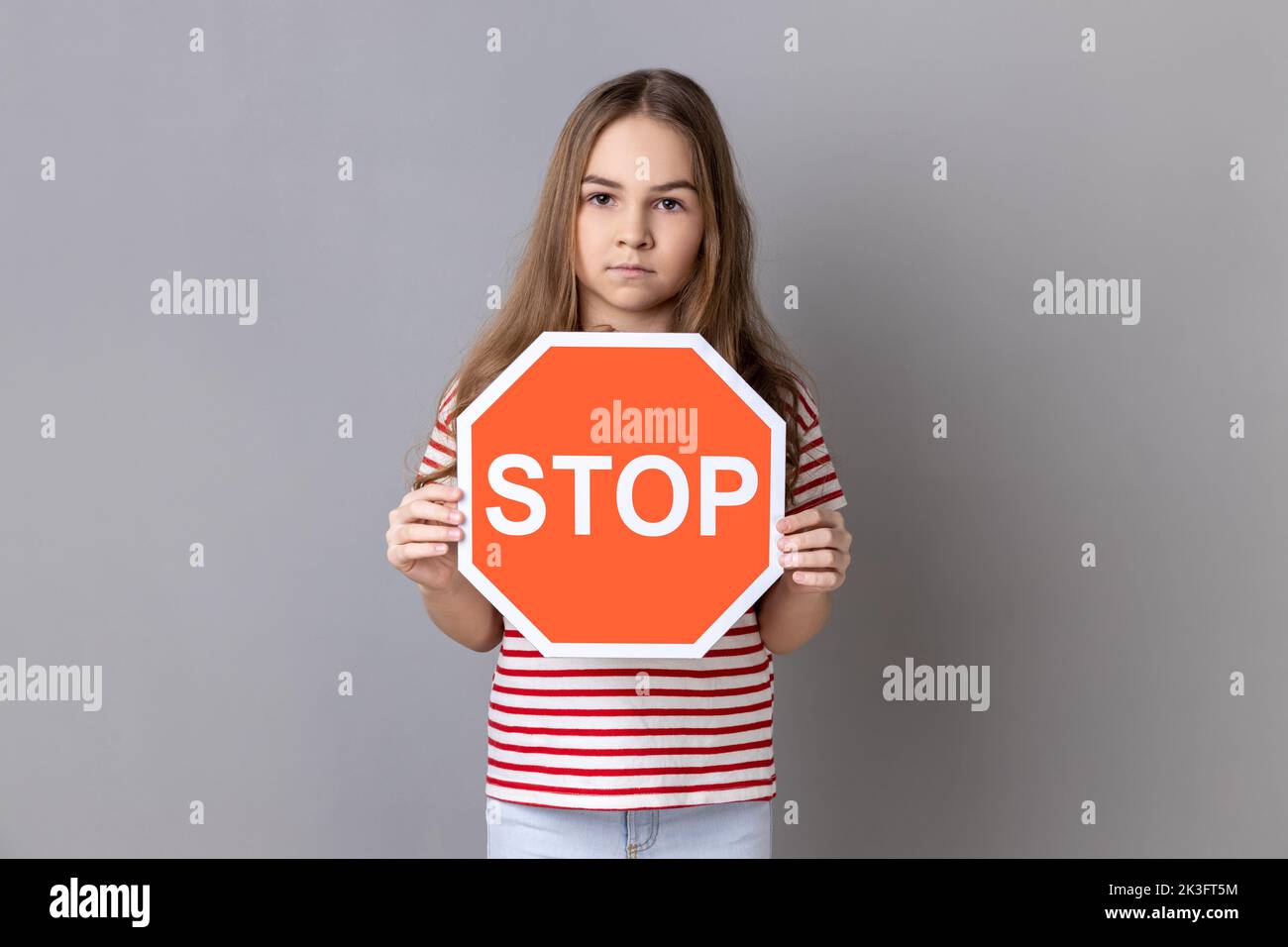 Portrait of serious strict little girl wearing striped T-shirt holding red stop sign, looking at camera with serious facial expression, prohibition. Indoor studio shot isolated on gray background. Stock Photo